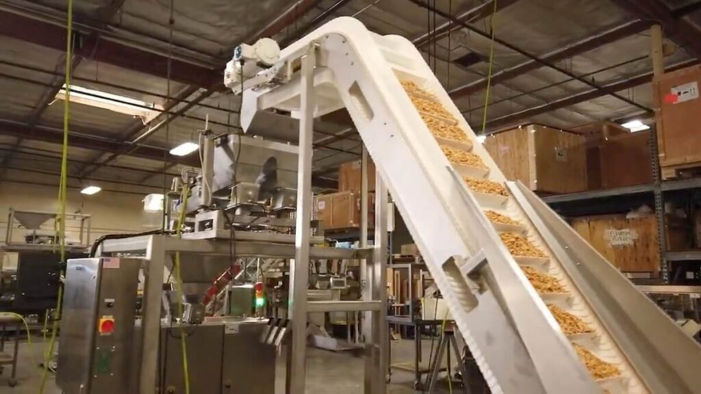 Industrial food processing facility featuring a vertical form-fill-seal machine and an inclined white cleated conveyor belt transporting dry food pieces (possibly cereal or snacks) upward for packaging. The setup includes stainless steel equipment, control panels, and is located in a warehouse with crates and shelving in the background.