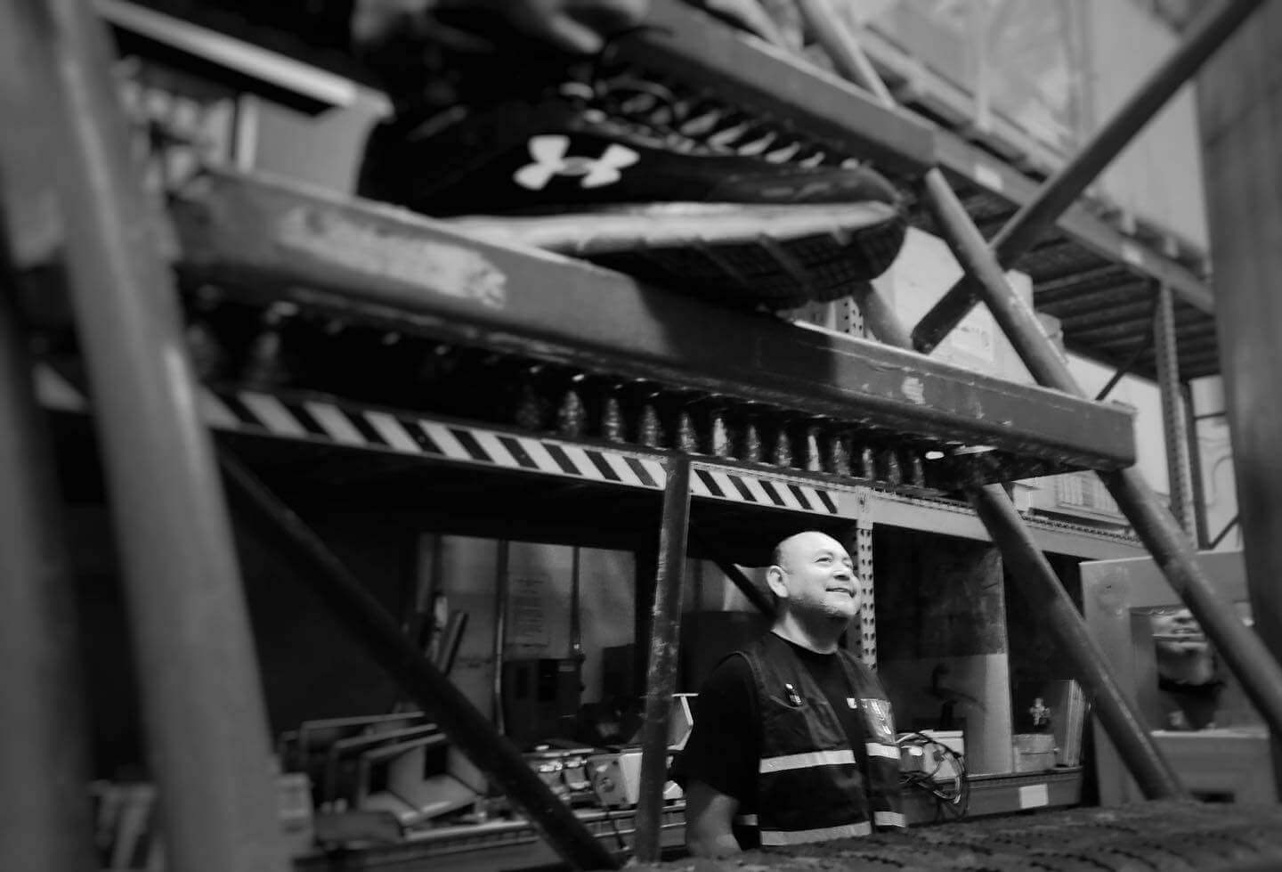 Black-and-white photo of a smiling man wearing a reflective safety vest, standing in an industrial warehouse beneath a shelving unit. The shelves above him hold equipment, including a pair of Under Armour shoes. Various tools and components are stored on the shelves behind him. Diagonal metal bars frame the scene, suggesting structural support beams.