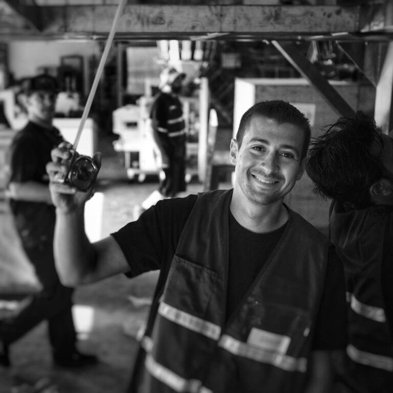 Black-and-white photo of a smiling worker in a reflective safety vest holding a tape measure in a warehouse or industrial setting. The background includes other workers and machinery in soft focus, adding context to the busy, productive environment.